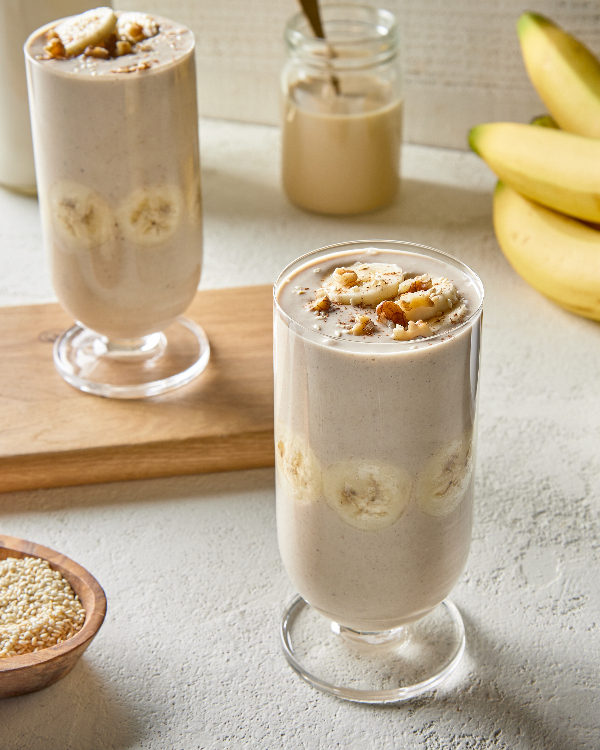 Deux grands verres de smoothie à la banane, avoine et tahini, l'un sur une planche à découper en bois, présentés avec un bol de graines de sésame, un pot de boisson à base de lait d'avoine et une grappe de bananes.