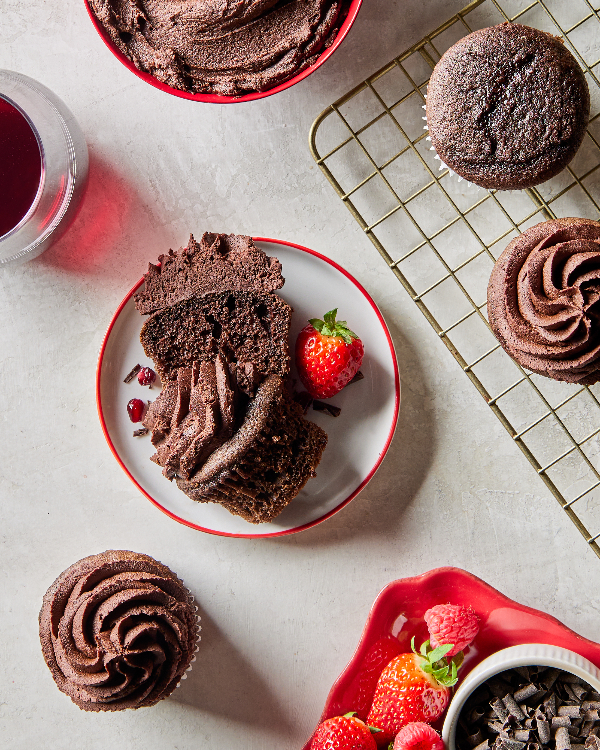 Cupcakes au chocolat et au vin rouge, certains natures, d'autres garnis de ganache, avec deux sur une grille de refroidissement, un sur un comptoir et un coupé en deux dans une assiette, présentés avec un bol de ganache, de chocolat râpé et de fraises.