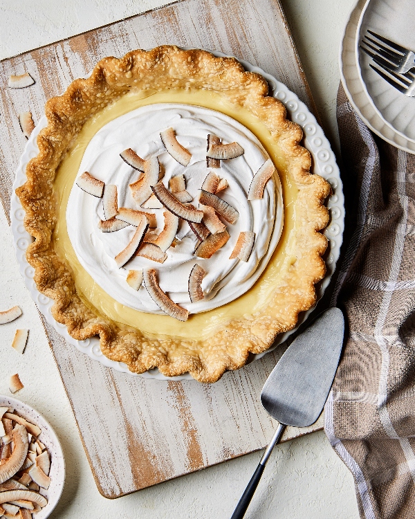 Tarte à la crème de coco fraîchement cuite avec une croûte feuilletée dorée et un nappage crémeux, garnie de flocons de noix de coco grillés, servie sur un plat à tarte blanc classique, accompagnée d'une pelle à tarte sur une table en bois rustique.