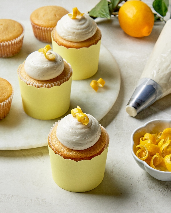 Trois cupcakes au citron avec un glaçage crémeux et un garniture de zeste de citron et trois autres sans garniture affichés sur un comptoir en marbre avec une poche à douille remplie et des citrons frais en arrière-plan.