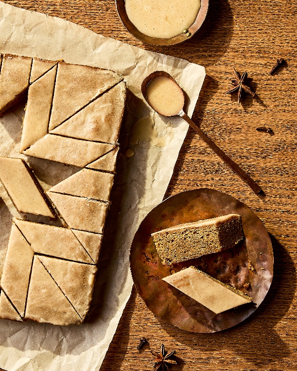 Des barres de gâteau épicées disposées sur du papier parchemin, coupées en losanges nets et recouvertes d'un glaçage brillant. Une assiette en métal contient une seule barre coupée en deux, mettant en valeur la mie tendre du gâteau. Une cuillère en bois avec du glaçage supplémentaire, un petit bol et des épices entières comme l'anis étoilé et les clous de girofle sont disposés à proximité, soulignant les saveurs chaudes et épicées du dessert