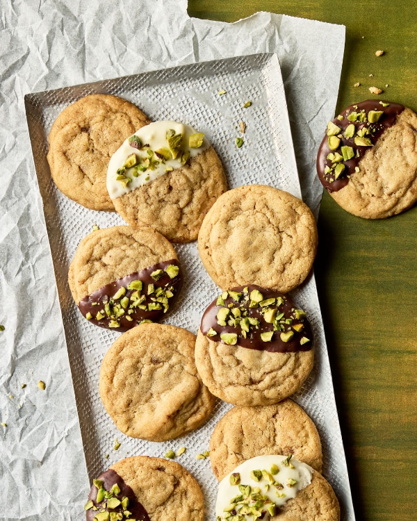 Biscuits sans pépites de chocolat disposés sur un plateau en métal texturé posé sur du papier parchemin. Certains biscuits ne sont pas décorés, tandis que d’autres sont à moitié trempés dans du chocolat blanc ou noir et saupoudrés de pistaches hachées. Un seul biscuit décoré repose sur la surface en bois vert à côté du plateau, avec des pistaches hachées éparpillées autour.