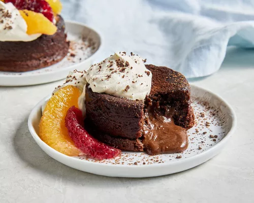 Deux gâteaux individuels au chocolat fondant garnis de crème fouettée et de cacao en poudre, agrémentés de segments d'orange et d'orange sanguine. L'un des gâteaux est montré avec un morceau manquant, laissant échapper une sauce au chocolat à partir du centre