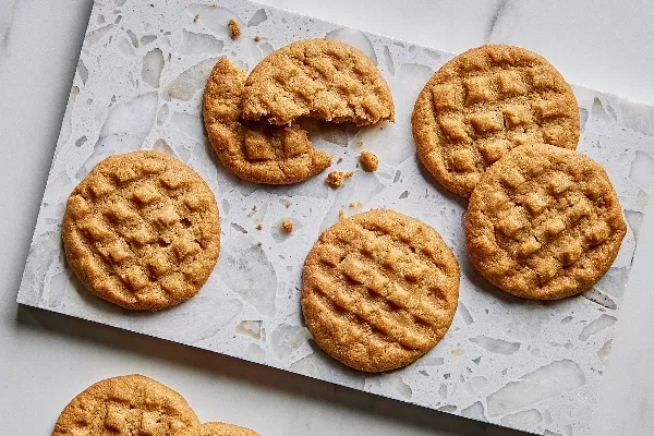 Sept biscuits au beurre d'arachide dorés avec des motifs distinctifs faits à la fourchette présentés sur du papier parchemin sur une surface en marbre, l'un partiellement mangé pour révéler une texture douce et moelleuse.