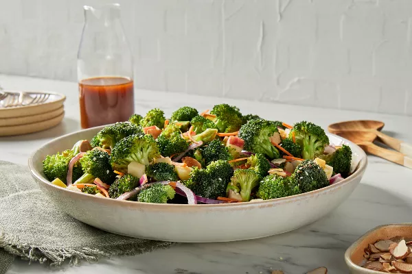 Un saladier de salade de brocoli avec une vinaigrette au gochujang est présenté sur un comptoir de cuisine, avec une carafe de vinaigrette, des assiettes et des cuillères de service en bois.