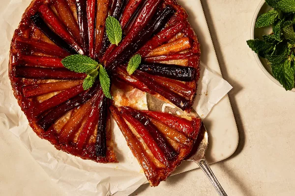 Une tarte tatin aux carottes et aux panais reposant sur du papier sulfurisé sur une planche à découper, présentée avec une tranche partiellement retirée à l'aide d'une pelle à tarte, garnie de feuilles de menthe fraîche.