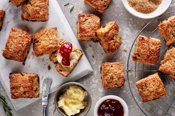Scones à la vanille, au romarin et aux dattes sur un plat et une grille de refroidissement, présentés avec des plats de beurre et de gelée et un bol de sucre turbinado.