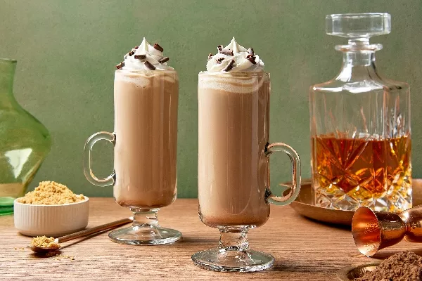 Deux grands verres de latte au whisky irlandais surmontés de crème fouettée et de copeaux de chocolat, présentés sur une table en bois avec un accompagnement de sucre brun, une carafe de whisky ambre en arrière-plan.