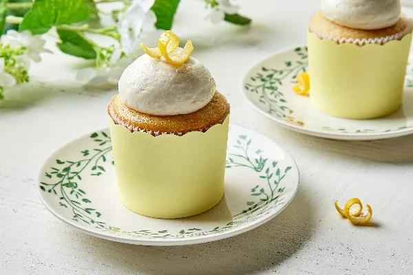 Des cupcakes au citron avec un glaçage à la vanille moelleux, ornés d'un zeste de citron torsadé, présentés sur une assiette à motif floral vintage posée sur une surface en bois blanc et devant un fond de verdure douce et de fleurs blanches pour une ambiance fraîche et printanière.