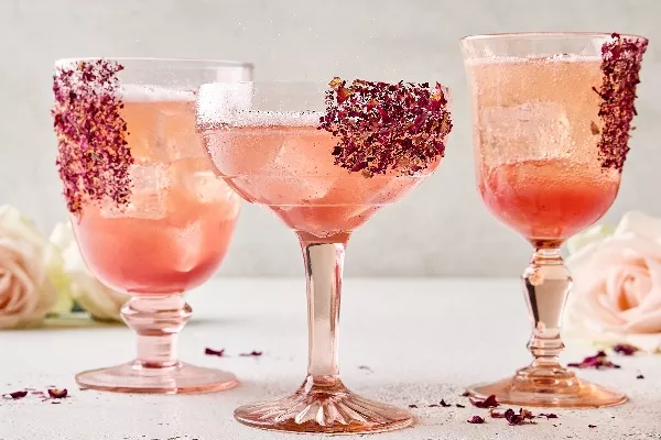 Trois verres à tige uniques de spritz litchi et rose sur glaçons. Des pétales de rose séchés garnissent les verres qui sont présentés dans un cadre neutre avec des roses fraîches en arrière-plan et des pétales de rose séchés dispersés autour.