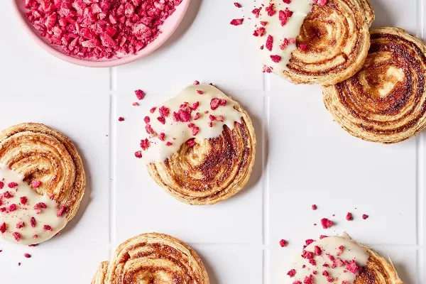 Vue aérienne de biscuits à la pâte feuilletée aux framboises avec glaçage au chocolat blanc et morceaux de framboises lyophilisées sur un comptoir carrelé blanc, avec un bol de framboises écrasées.