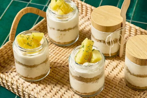 Un plateau en osier, adossé à un arrière-plan de carreaux verts, contient plusieurs verrines de cheesecake à l'ananas en portions individuelles. Chaque verrine présente des couches de gâteau au fromage crémeux et de croûte friable au graham cracker, garnies de morceaux d'ananas frais et d'une pincée de zeste de lime. Deux des verrines ont des couvercles en bois, et une d'entre elles est accompagnée d'une cuillère en bambou attachée
