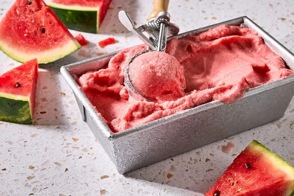 Un contenant en métal rempli de sorbet crémeux à la pastèque, en train d'être portionné avec une cuillère à crème glacée vintage. Des tranches de pastèque fraîche sont dispersées autour du contenant sur un comptoir en calcaire de couleur claire.