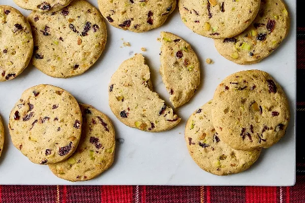 Des biscuits à la canneberge et à la pistache disposés sur une surface de marbre blanc. Les biscuits tranchés et cuits au four présentent des morceaux visibles de canneberges séchées et de pistaches, ajoutant texture et couleur. Un biscuit est coupé en deux, révélant une mie tendre. Un tissu à carreaux rouges ajoute une touche festive au bas de l'image.