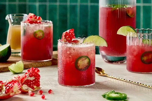 Trois verres de Fizz Épicé à la Grenade et au Gingembre, d'une couleur rouge vibrante avec des tranches de jalapeño frais. Les boissons sont garnies de quartiers de lime et de graines de grenade. Une carafe remplie de la même boisson se trouve en arrière-plan, aux côtés de quartiers de lime, de morceaux de grenade et d'une petite carafe de sirop de gingembre. La scène se déroule sur un fond de tuiles vert émeraude.