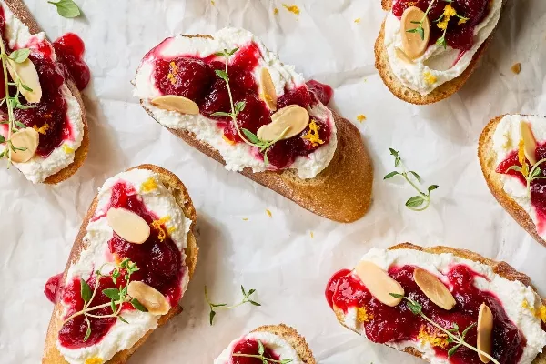 Un gros plan sur plusieurs crostinis garnis de feta fouettée, de chutney de canneberges, d'amandes effilées et de thym frais.