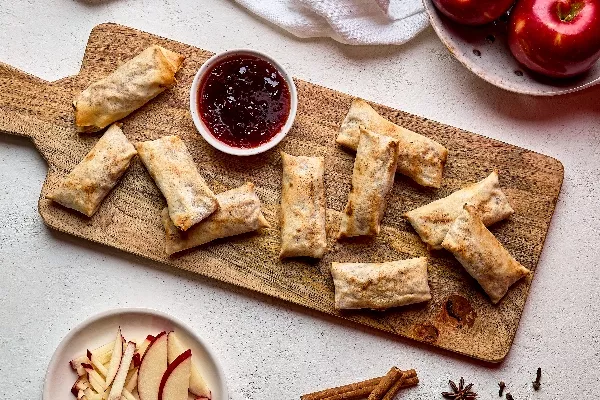 Rouleaux de printemps croustillants aux pommes disposés sur une planche de service en bois avec un petit bol de sauce rouge au centre. Des pommes rouges fraîches et des bâtonnets de pommes tranchés sont disposés à proximité, ainsi que des bâtons de cannelle, de l'anis étoilé et des clous de girofle.