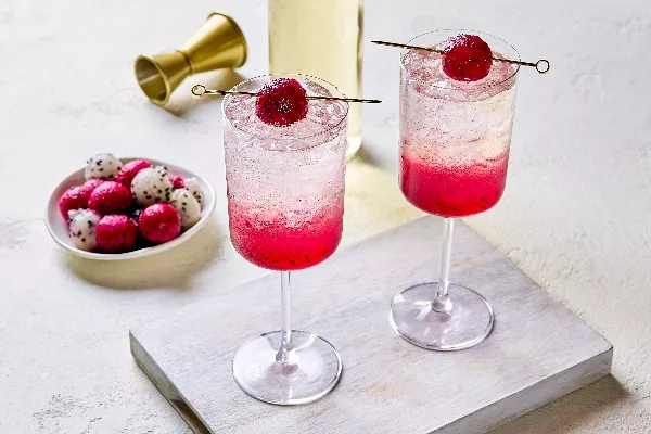 Deux verres de spritz à la pitayaha servi sur glace, avec un dégradé de rose provenant des morceaux de fruit au fond. Chaque verre est garni d’une boule de pitayaha sur brochette. Une assiette remplie de boules de pitayaha rouge et blanche, un doseur à cocktail doré et une bouteille sont placés en arrière-plan.