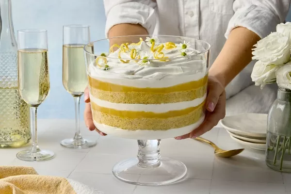 Deux mains qui tiennent un bol de service en verre rempli d’une bagatelle au citron et à la fleur de sureau avec ses couches de gâteau éponge, de curd au citron et de crème fouettée. La bagatelle est couronnée de rubans de zeste de citron et de délicates fleurs blanches. Des flûtes à champagne, une bouteille et des roses blanches dans un vase en verre sont visibles en arrière-plan.