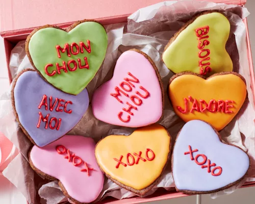 Des biscuits en sucre en forme de cœur décorés de glaçage royal avec des messages romantiques écrits dans le glaçage, présentés sur des carreaux blancs avec des bols de glaçage royal et une poche à douille.