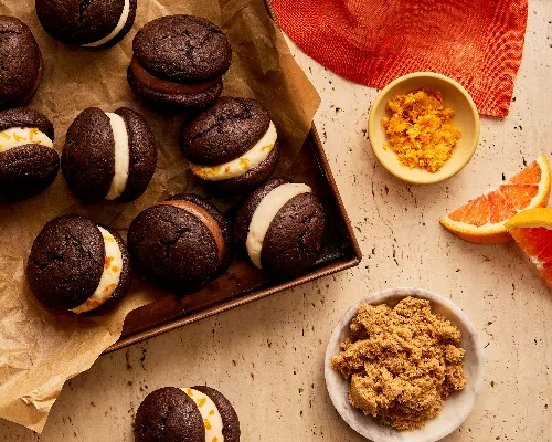 Des mini whoopie pies au chocolat avec diverses garnitures sur une plaque de cuisson doublée de papier parchemin, présentés avec un bol de sucre brun foncé, un bol de zeste d'orange et des quartiers d'orange